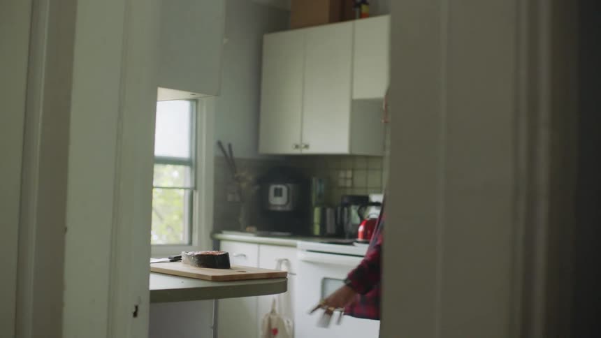 A man is standing in a kitchen holding a knife over a piece of meat on a cutting board and he puts down the knife and walks away