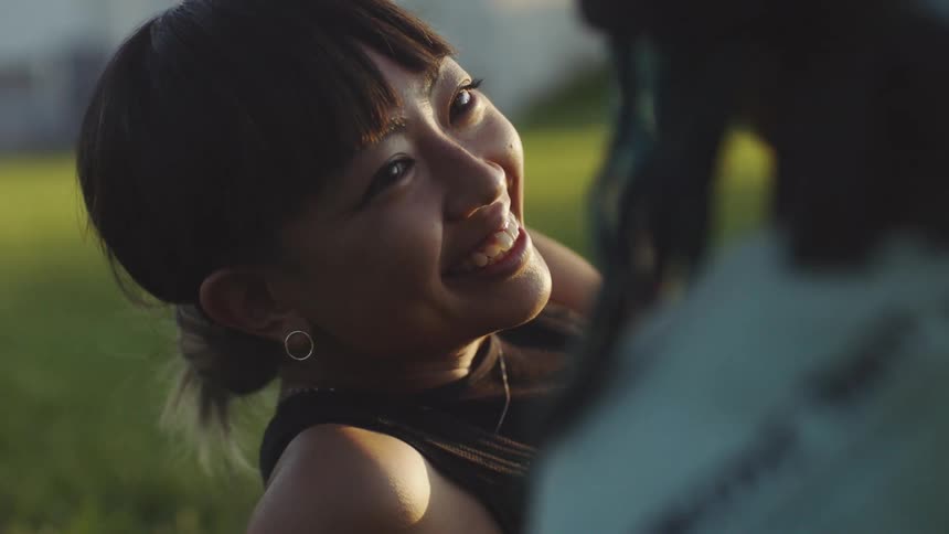 Woman with earring with bun hair and sleeveless brown shirt talks to man with braids lying down on grass in public park