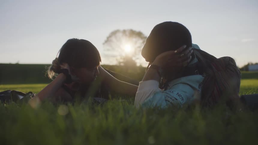 Couple lie on their sides on lawn with their head propped up on their arms looking at each other on sunny day