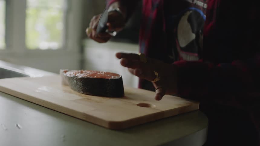 A man walks up to a cutting board at home and picks up a knife an is about to cut a piece of fish but then puts the knife down and backs up