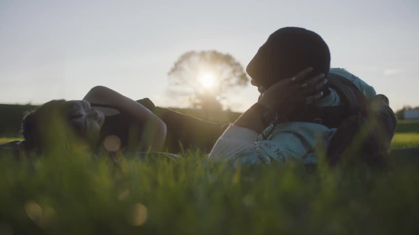 A teenage boy and girl lie in the grass talking to one another and they both raise a leg in the air and touch their toe
