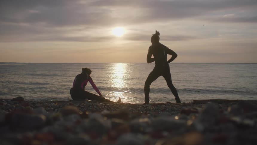 A man and woman stretch while standing on rocks along the coast