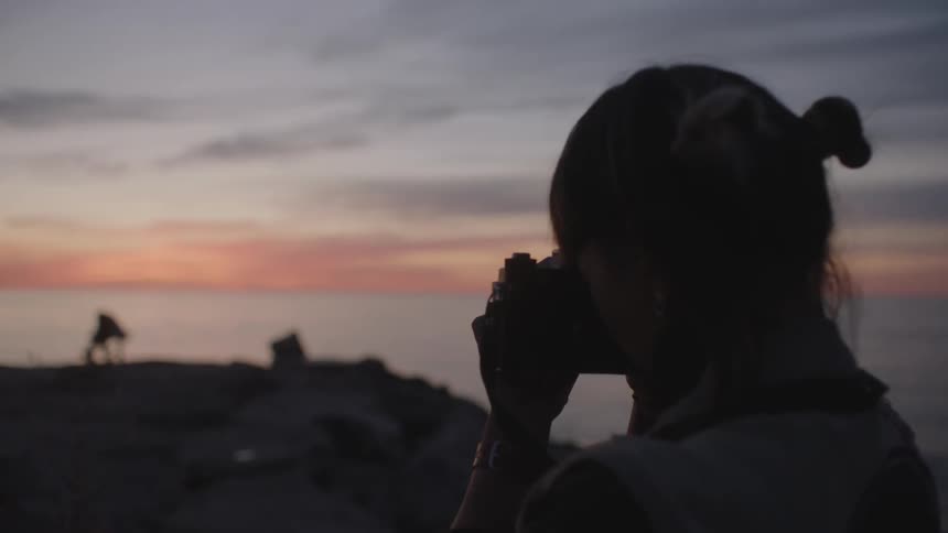 The silhouette of a woman photographing a person doing yoga near the ocean at sunset