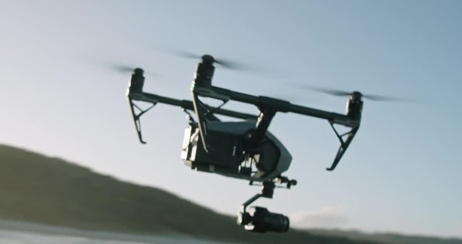 A remote controlled drone with a camera mounted beneath it flies along a rocky coastline during the day