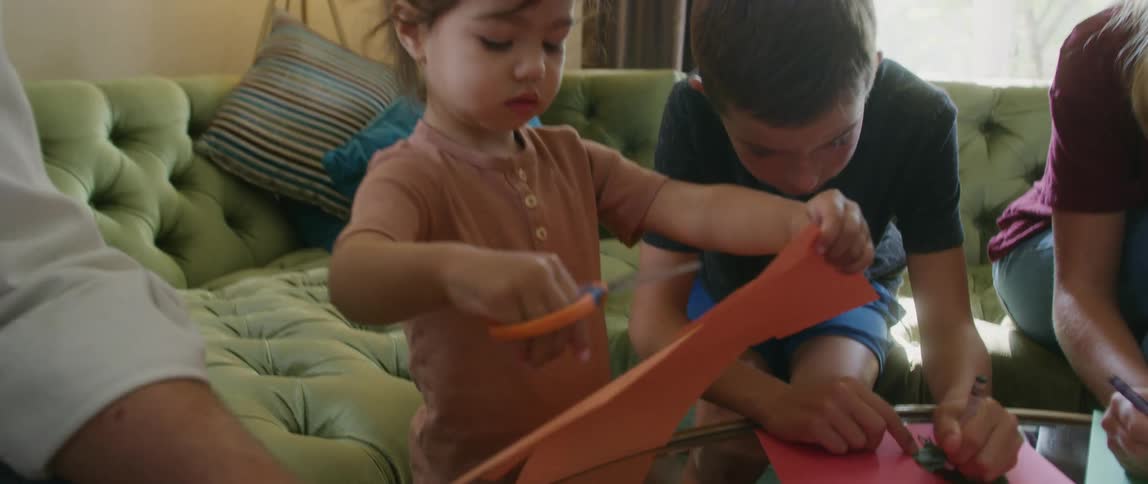A toddler uses scissors to cut construction paper while crafting with her family