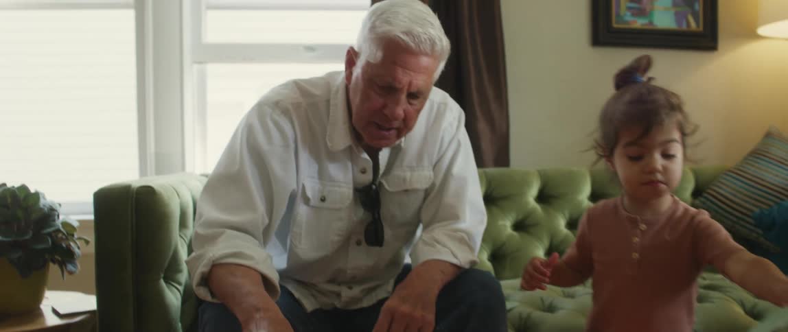 An elderly man sits on a couch and watches as children cut shapes out of colored paper