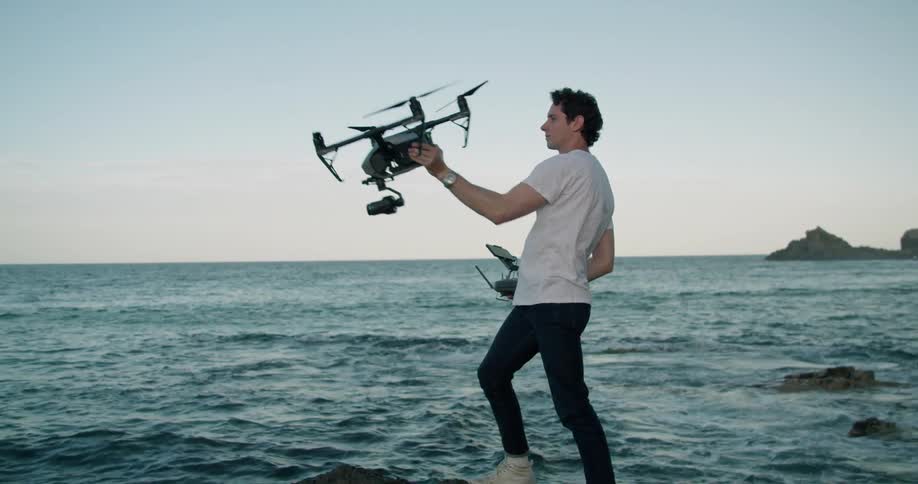 A man stands on the shore and reaches up and takes his drone out of the air