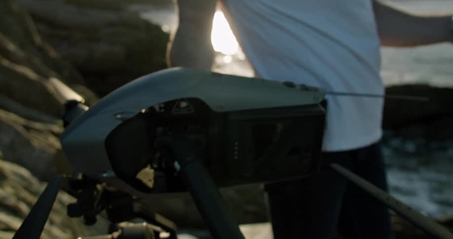 A man is standing near the water holding a drone in one hand and the remote controller in the other hand