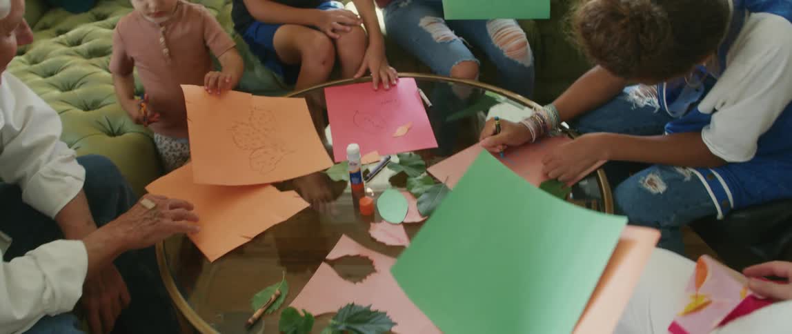 An elderly woman picks up some colored paper and walks over to a couch where her grandchildren and husband are sitting and drawing and cutting the paper and hands them more paper