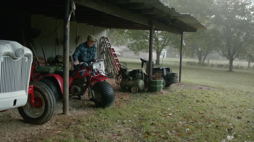 An elderly man is testing the headlight of an all terrain trike under an awning next to a shed on a rainy day