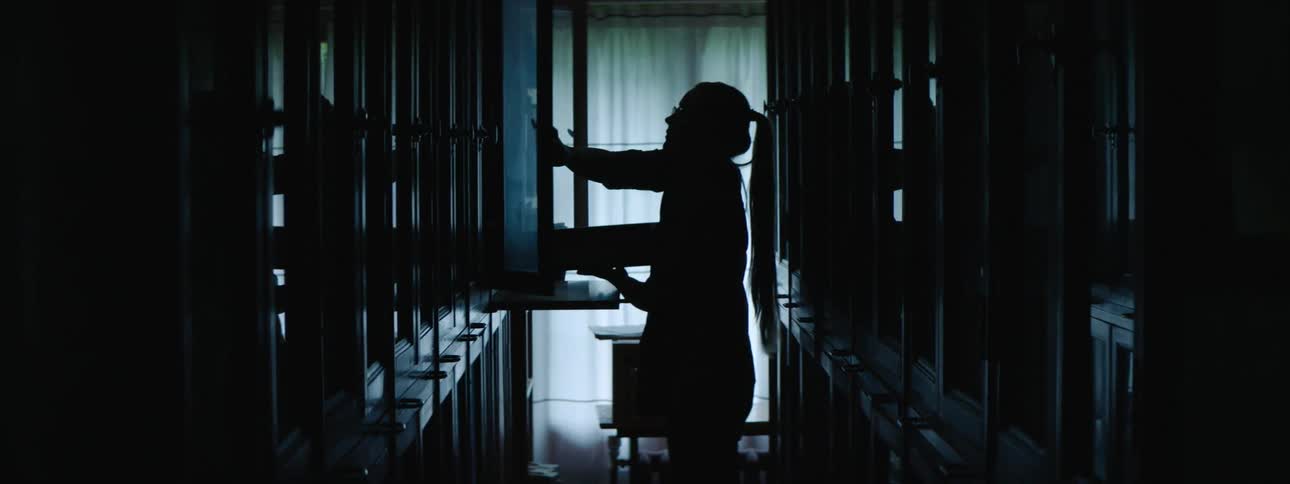 A woman walks in a dimly lit hallway lined with cabinets and puts a box in one and then closes the door