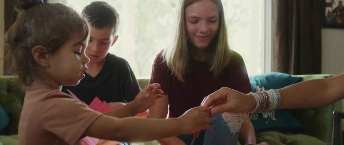 Four children sit at a couch with an elderly man as they all cuts shapes out of colored paper