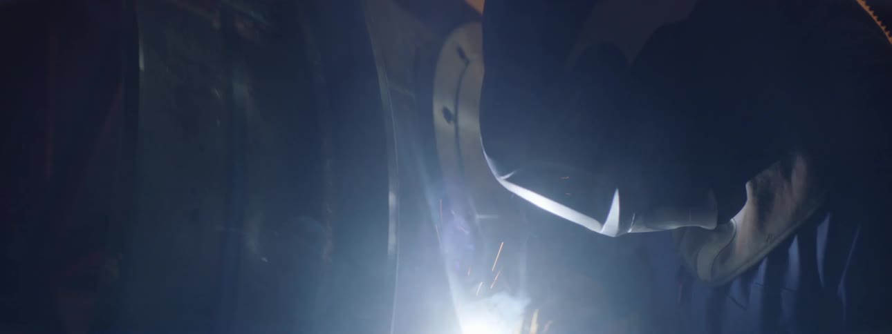 Person wearing coveralls and protective face shield welds metal framework in workshop