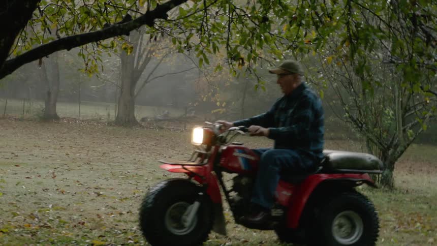 A man is riding a three wheel all terrain vehicle across a yard with many trees