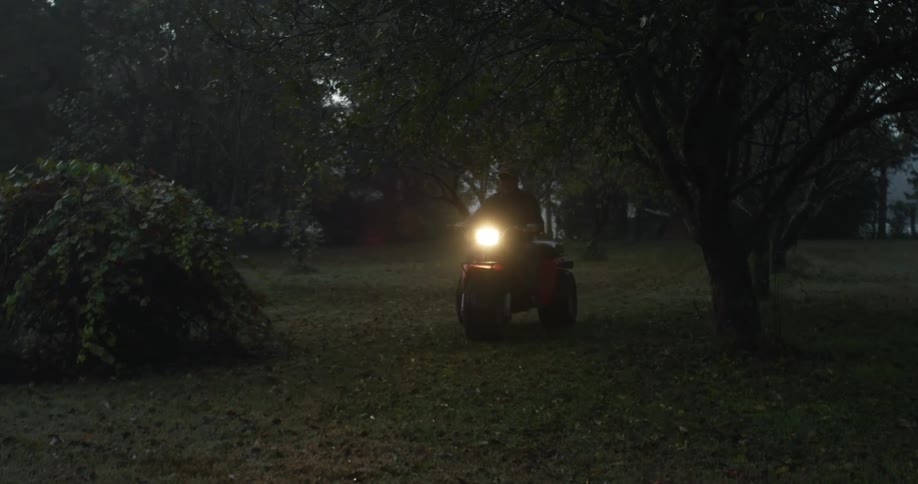 A man is riding a three wheel all terrain vehicle with the headlight on across a yard with many trees