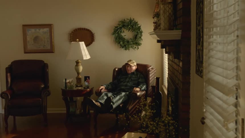 An elderly man sits in a chair at home with his feet up