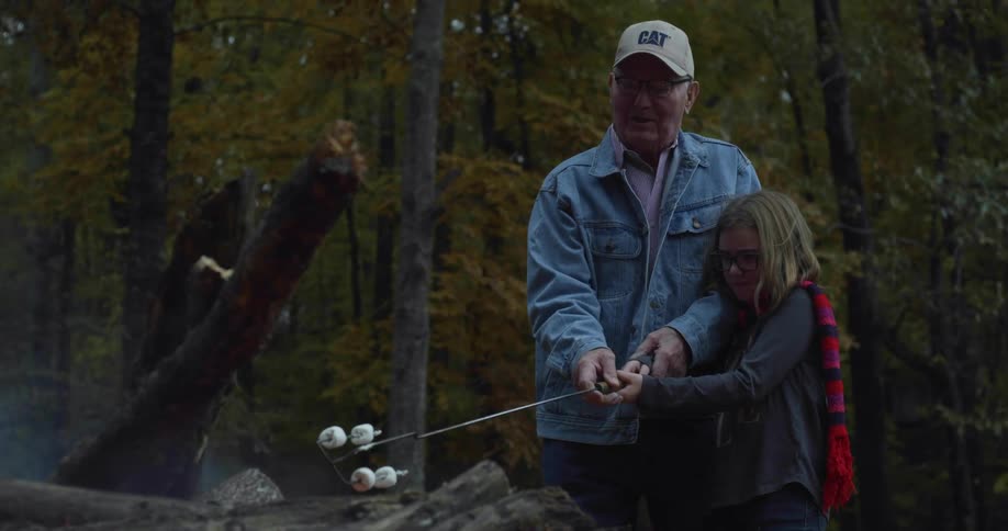 A grandfather helps his granddaughter to grill marshmallows over a campfire in a forest during the day