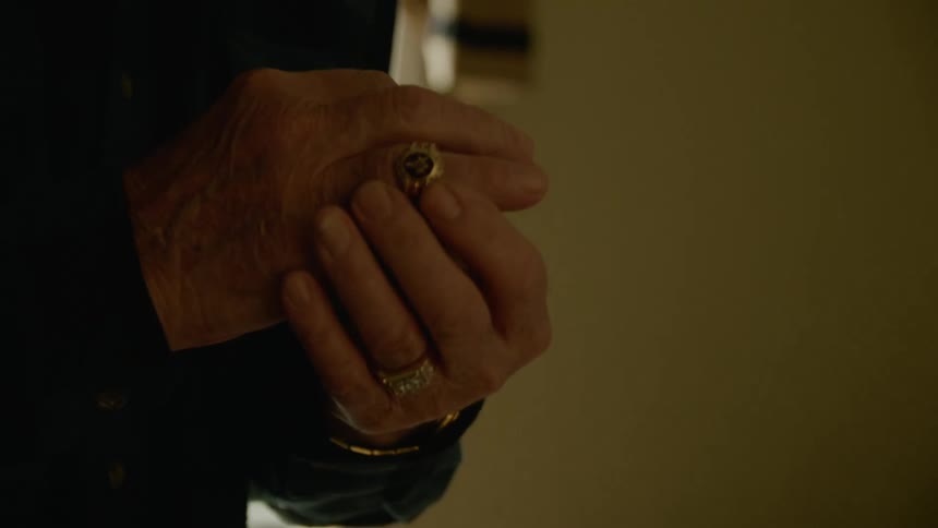An elderly man picks a ring up off of a bed and slips it on his finger