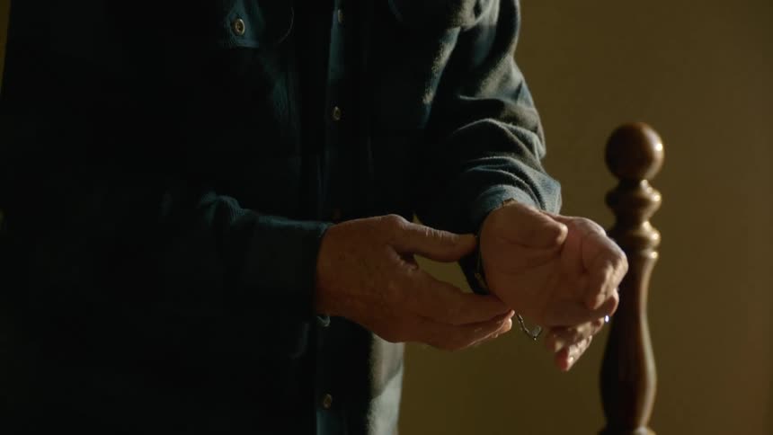 Elderly man with cap glasses and dark blue plaid shirt wears golden wristwatch in bedroom