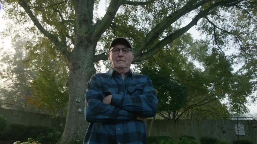 An elderly man stands in his yard with his arms crossed