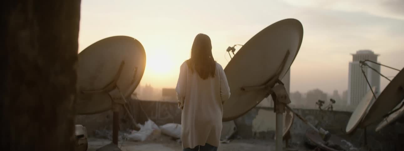 A woman walks out onto a balcony with satellite dishes overlooking a city and takes a picture of a building