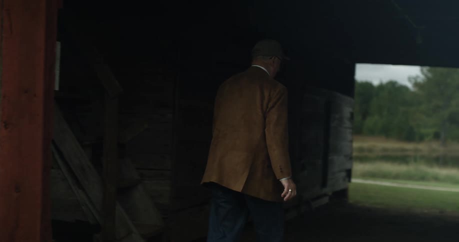 A man wearing a cap and blazer walks past the equipment barn and pauses at a door