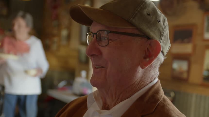 An elderly man wearing a cap is standing inside a restaurant talking to someone and smiling and laughing as a woman in the distance carries food