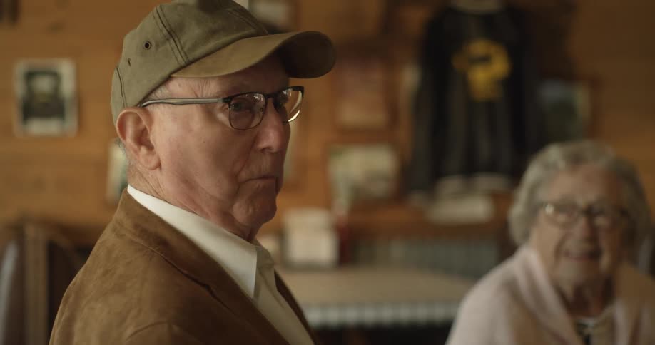 An elderly man wearing glasses and a cap is standing inside a restaurant talking to someone and an old woman is sitting nearby