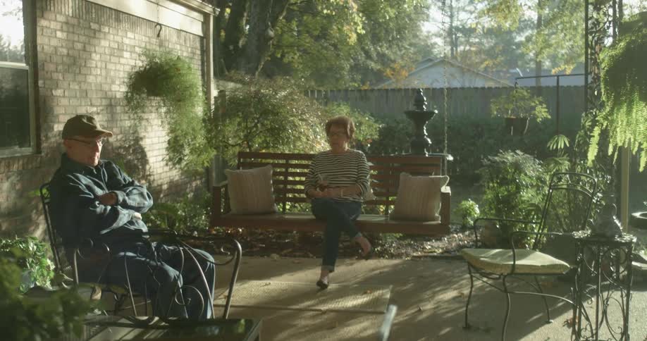 A man sits in a chair in his backyard while his wife sits nearby on a bench swing