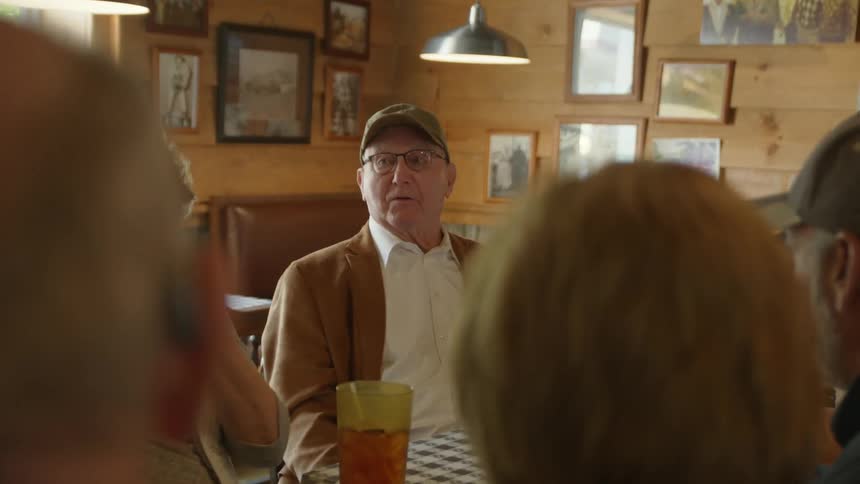 A group of elderly people sit at a table in a restaurant and talk as a woman is served a meal
