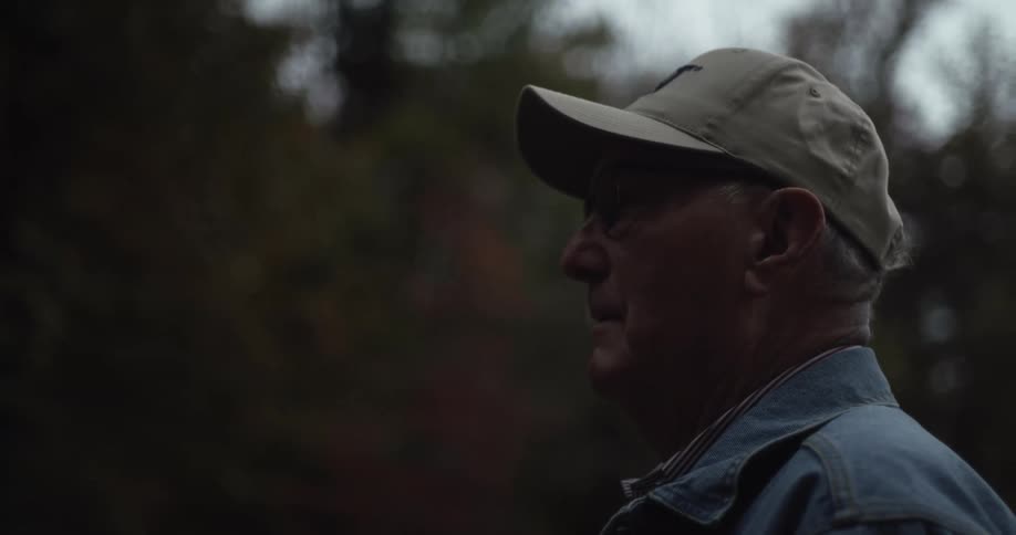Older man stands outdoors wearing a baseball cap and a denim jacket