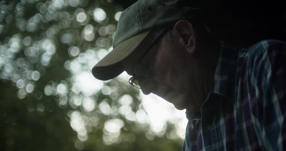An elderly man wearing glasses and a cap is standing outside looking down at something
