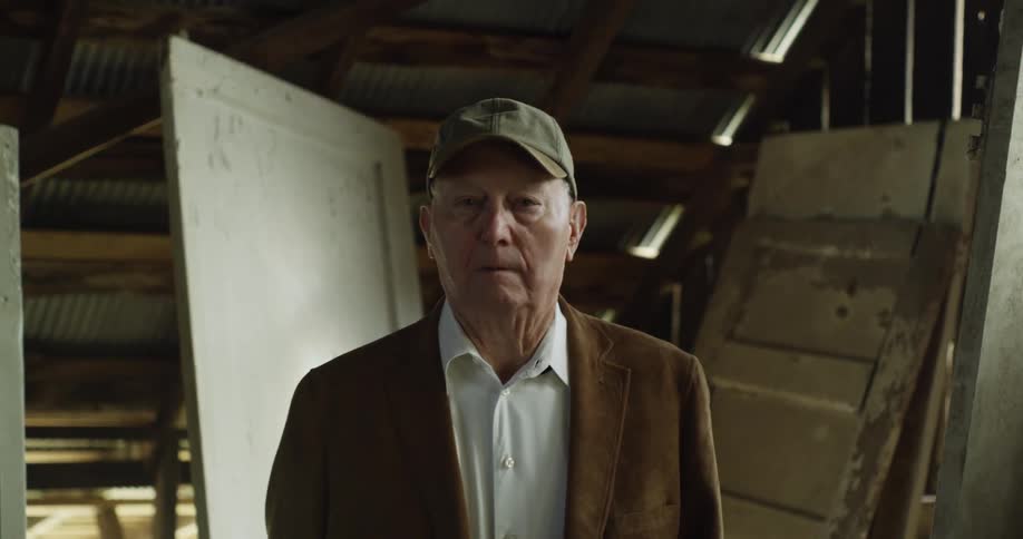 Elderly man with cap white shirt and brown blazer jacket looks at camera in the attic
