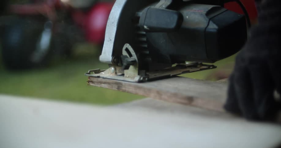 Gloved hands brace a plank of weathered wood as it is cut using a power hand saw