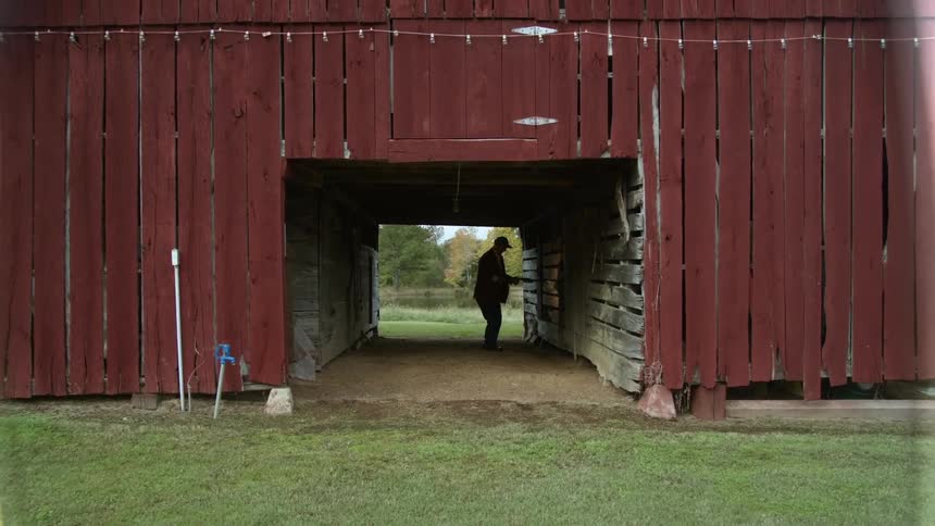 A man walking to his barn pauses to bend and pickup a piece of metal and replace it