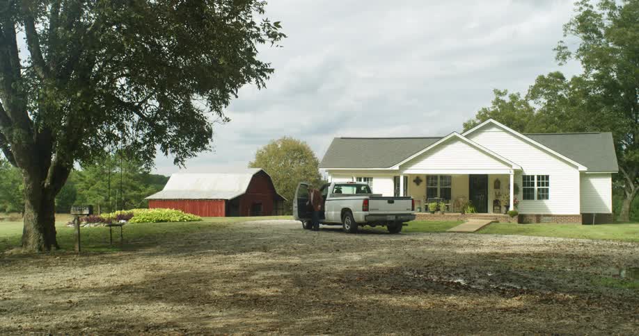 A pickup truck pulls up in front of a farmhouse and a man gets out walking towards the red barn