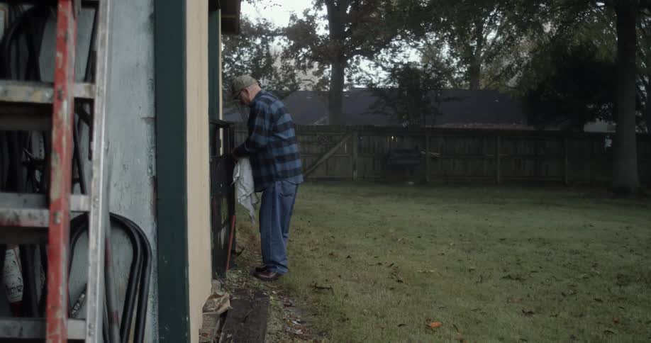 Elderly man with cap glasses blue plaid shirt and jeans walks in backyard carrying towels