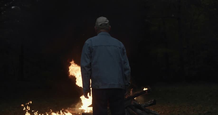 Man stands in field near forest watching burning bonfire