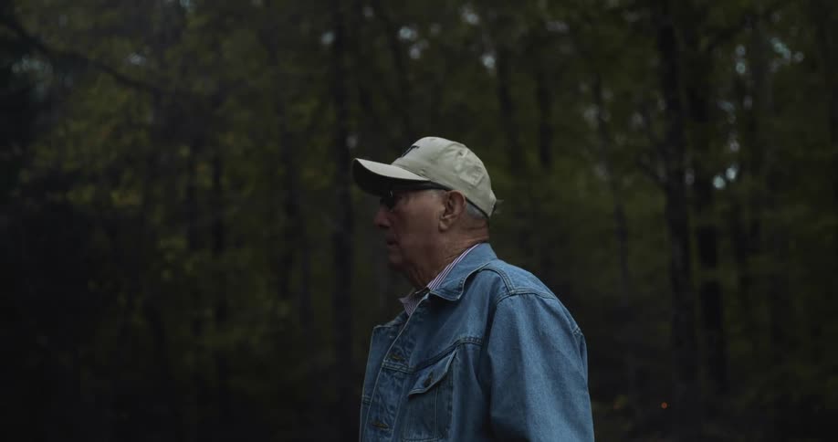 An elderly man is standing and looking around while in a woodland area during the day