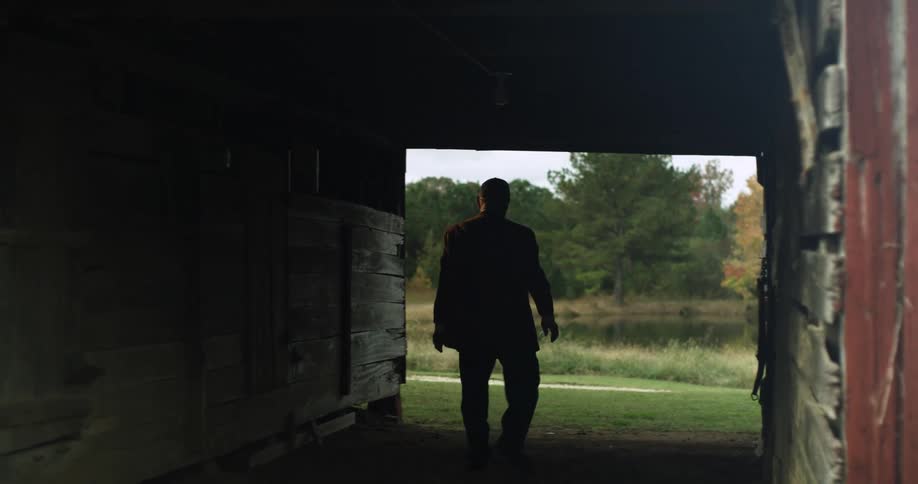 An elderly man hangs a tool on the inside wall of a barn and walks out near a pond