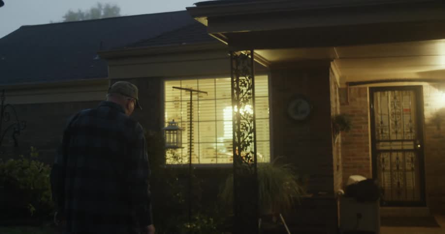 An elderly man walks towards a home and takes a birdhouse down that is hanging out front