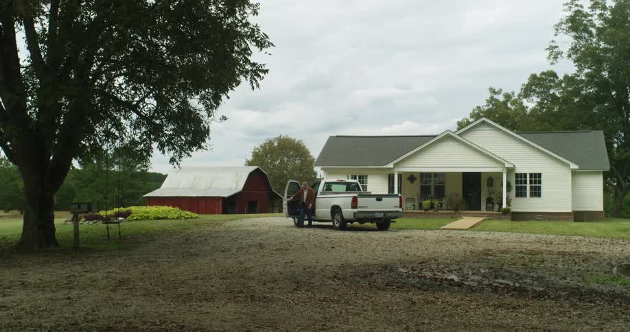 A man pulls his truck up in front of a house and gets out and starts to walk towards the barn