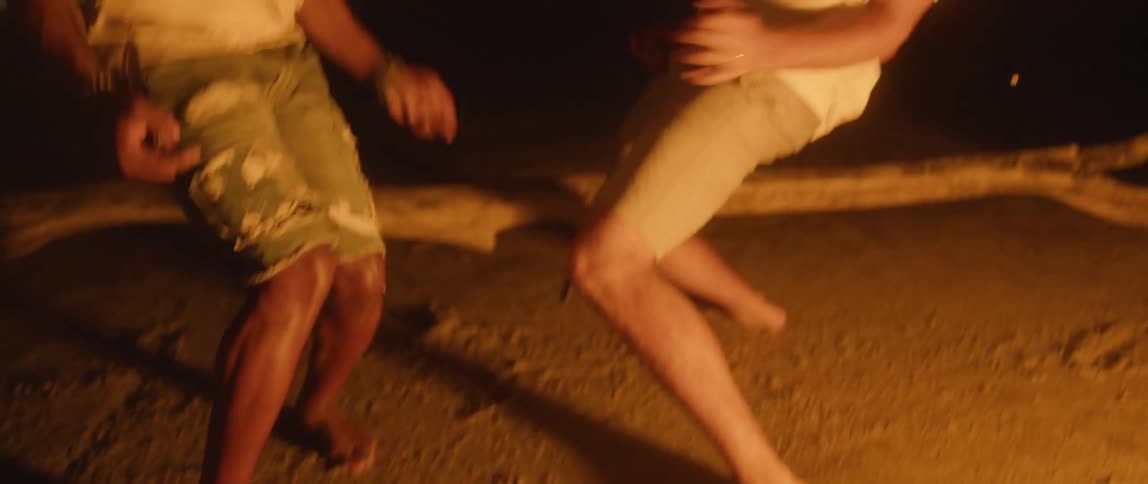 Two men dance facing each other laughing and smiling as flames from the campfire glow