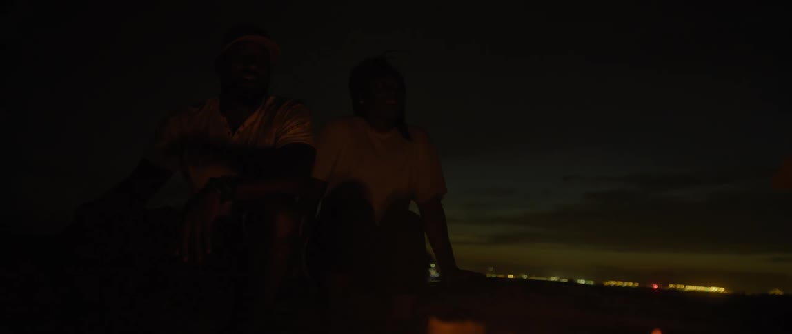 A man and woman sit together next to a campfire on the outskirts of a city at night