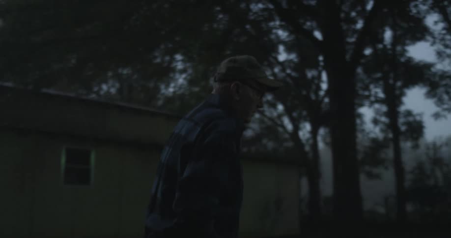 Elderly man with cap glasses and plaid shirt walks in backyard at dawn