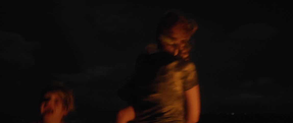 A woman her holds toddler daughter as they dance with her young son around a campfire