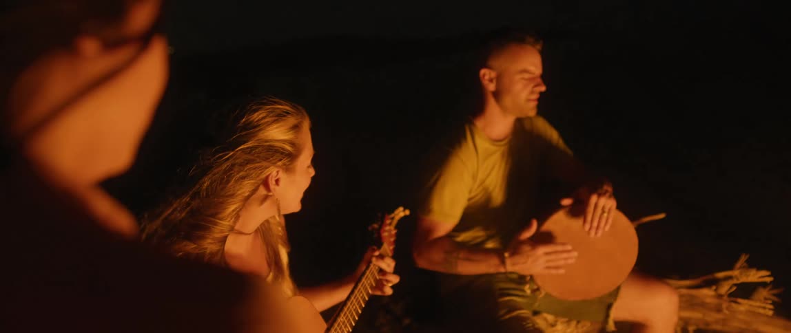People are sitting at a campfire at night and playing music on a guitar and hand drum