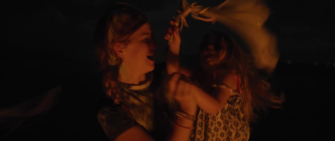 A mother holds a young girl in her arms and they dance and around and spin in circles as the girl waves a flag in the night air