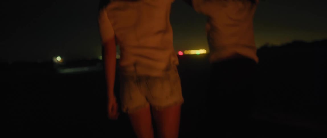Two women are dancing together near a campfire on a beach at night with city lights glowing in the distance