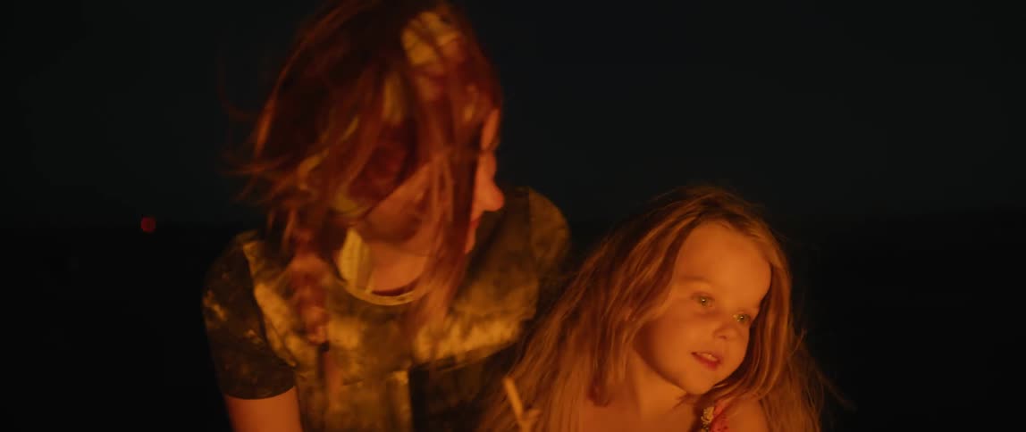 A mother sits beside her daughter and smiles as they look in to a fire burning at night